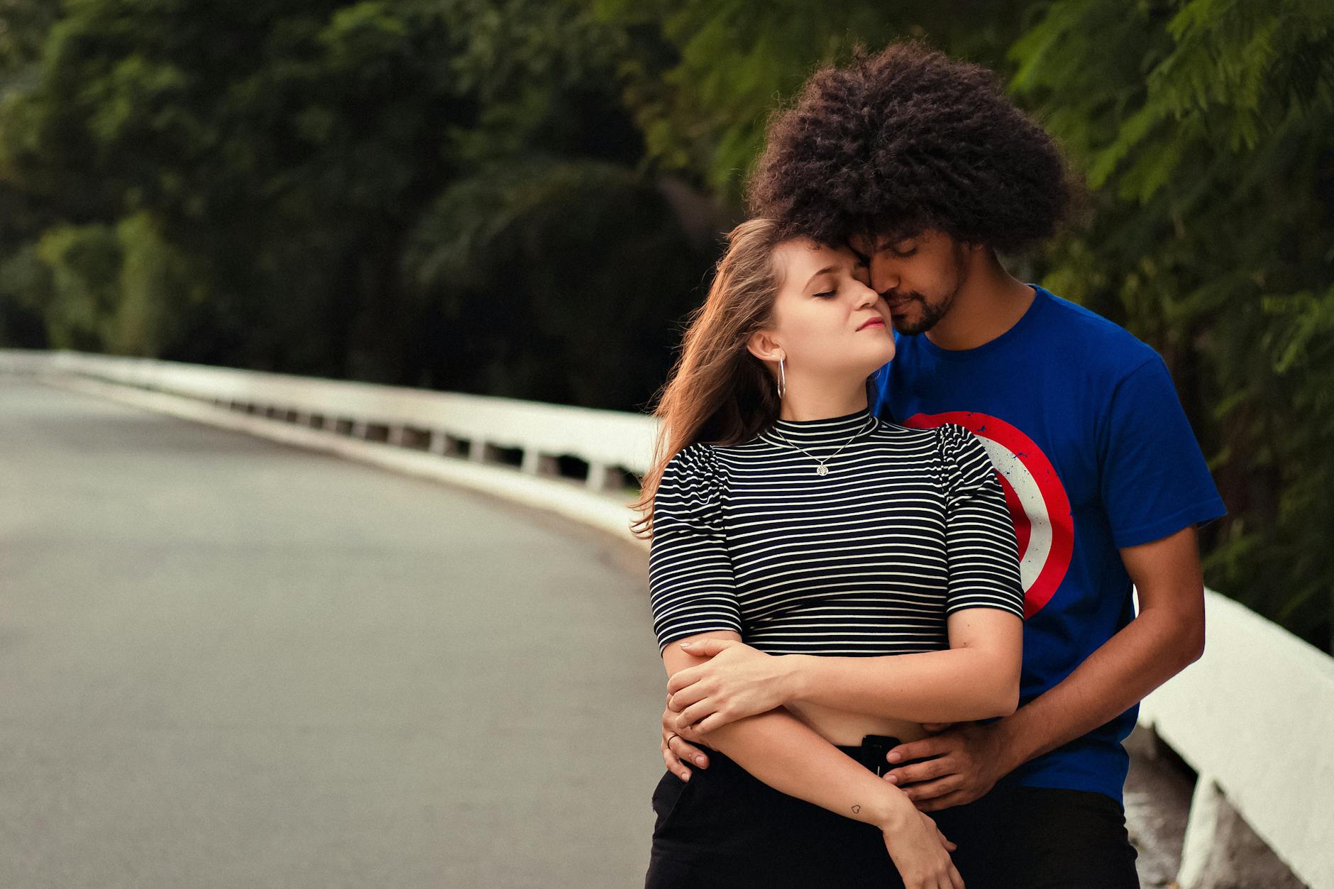 Loving multiracial boyfriend and girlfriend cuddling on asphalt road surrounded with green trees